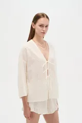 Blusa de algodón blanco con diseño de broderie, cuello en V y cierre con lazos frontales, mangas largas de corte holgado.