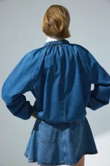 Campera bomber oversize de denim azul, con cuello elevado fruncido, cierre frontal y bolsillos laterales. Cuenta con mangas raglán y elásticos en puños y bajo.