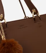 Cartera estilo satchel de tamaño mediano, confeccionada en poliuretano color marrón con textura tipo cuero. Cuenta con asas de mano reforzadas, correa larga desmontable, cierre superior de cremallera y un colgante decorativo de pompón de pelo sintético.