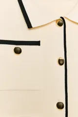 Chaqueta de punto color crema con ribetes en contraste negro, cuello solapa, manga larga y cierre frontal con botones dorados. Presenta bolsillos de plastrón en el delantero con detalle de botones.
