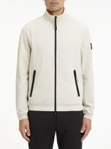 Campera Calvin Klein color beige claro, confeccionada en nailon arrugado reciclado. Tiene cuello alto, puños y bajo elásticos, cierre con cremallera, bolsillos con cremallera en la cintura, abertura de ventilación en la espalda, forro de poliéster reciclado e insignia con el logo de goma de Calvin Klein en la manga.