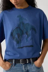 Remera de algodón color azul con estampa frontal de un cowboy montando un caballo, rodeado de pequeños puntos blancos. Presenta cuello redondo y mangas cortas con un corte holgado.