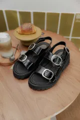 Sandalias de plataforma estilo chunky en color negro, confeccionadas en material sintético efecto charol. Presentan dos tiras ajustables con hebillas metálicas plateadas y una tira trasera con hebilla para mayor sujeción. Suela dentada de goma.