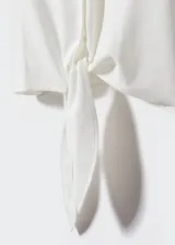 Camisa blanca de lino con cuello camisero, manga corta con vuelta, diseño crop y oversize. Tiene un bolsillo de parche en la parte delantera, cierre de botones y lazo ajustable.