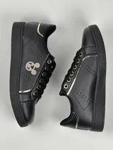 Zapatillas deportivas de cuero sintético en color blanco, beige y negro, con cordones y aplique decorativo dorado con forma de moño.