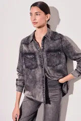 Camisa de denim celeste con cuello clásico, cierre frontal con botones a presión, dos bolsillos con solapa abotonada en el pecho y mangas largas con puños abotonados.