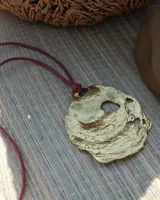 Collar artesanal con dije central de metal con textura orgánica irregular que evoca una ostra. El dije está montado sobre un cordón de algodón natural, permitiendo un uso versátil y regulable.