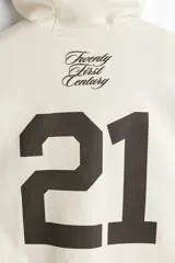 Canguro de algodón pesado color beige, con capucha de doble capa, hombros caídos y bolsillo canguro. Puños y bajo en punto acanalado. Estampado frontal con el número "21" en color negro.