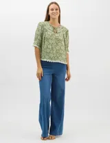Blusa de gasa estampada con motivos botánicos en color verde, con escote redondo, abertura frontal con lazos de ajuste y mangas cortas. Presenta detalles de encaje en el escote, mangas y ruedo.