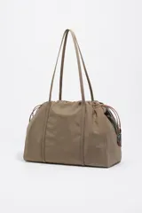 Bolso de hombro Bimba y Lola de tamaño grande, confeccionado en nylon negro con estructura blanda. Presenta dos asas largas, cierre superior y detalles de cordones laterales en tonos tierra. Incluye un parche frontal con el logo de la marca grabado.