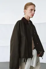 Chaqueta encerada de corte oversize, color marrón oscuro, con cuello de pana en contraste. Presenta cierre frontal con cremallera y botones a presión ocultos bajo solapa, bolsillos delanteros y mangas largas con puños ajustables.