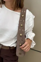 Cartera de hombro confeccionada en gamuza color habano, con diseño de correa ancha decorada con tachas metálicas circulares. Presenta cierre superior y placa metálica con logo en el frente.
