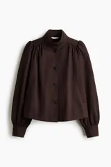 Campera corta de sarga color marrón oscuro, con cuello alto, cierre frontal con botones y bolsillos laterales. Presenta mangas largas abullonadas con fruncidos en los hombros y puños ajustados.