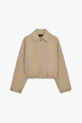 Chaqueta bomber color beige con cuello solapa, manga larga y falsos bolsillos delanteros. Presenta acabados abullonados y elásticos, y cierre frontal con cremallera.