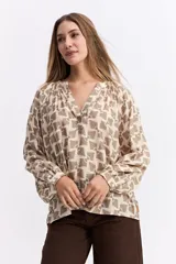 Blusa de corte relajado con estampado geométrico en tonos beige y marrón. Presenta escote en V con detalle de solapa, mangas largas abullonadas con puños abotonados y una caída liviana.