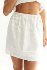 Falda corta blanca de lino, con cintura elástica y sección superior plisada. Presenta capas superpuestas en el ruedo.
