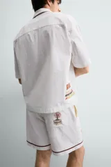 Bermuda de corte relajado confeccionada en algodón, color blanco con vivo en contraste color marrón en el ruedo. Presenta cintura elástica con cordón ajustable y un bordado decorativo en la pierna derecha que incluye una palmera y un gallo.