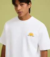 Remera de algodón blanca, corte relajado (relaxed fit), con cuello redondo y manga corta. Presenta un pequeño estampado frontal en el pecho con un sol estilizado y el texto "Sun Settings".