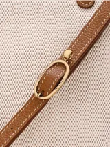 Bolso de mano pequeño con diseño estructurado, confeccionado en lona color beige con bolsillo frontal redondeado en cuero color marrón. Cuenta con asas cortas superiores y herrajes metálicos.