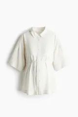 Camisa de viscosa color marfil, de corte holgado con hombros caidos, mangas cortas hasta el codo, cuello camisero y botonadura frontal. Incluye un cinturon fino extraible para ajustar en la cintura y aberturas laterales en el bajo.