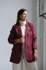 Blazer de corte sastrero en color bordó, con solapas clásicas, bolsillos frontales con solapa y cierre frontal con botones. Presenta una textura sutil en la tela y un broche decorativo en forma de sol en la solapa.