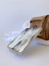 Cinto de cuero color marrón con forro de cuero y hebilla rectangular con diseño de guampa en blanco y negro con marco metálico plateado.