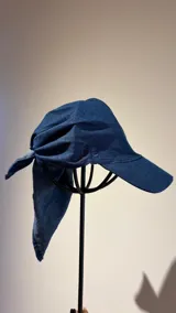 Gorro tipo pañuelo con visera rígida azul marino, confeccionado en lino con estampado de rayas grises y blancas. Presenta un diseño envolvente con caída trasera y detalle de logo bordado en el lateral.