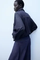 Cazadora bomber de denim oscuro con cuello subido, manga larga y bolsillos frontales con solapa. Presenta cierre frontal de cremallera y bajo elástico.