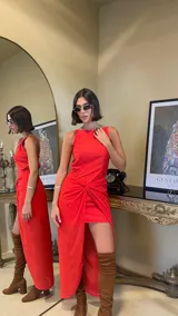 Vestido largo rojo con diseño asimétrico, escote redondo y detalle de nudo drapeado en la cintura que crea una abertura frontal, dejando ver una falda corta interior.