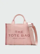 Bolso tipo tote bag de Marc Jacobs en color beige, confeccionado en lona resistente. Presenta el texto "THE TOTE BAG" y "MARC JACOBS" impreso en la parte frontal en color negro. Cuenta con asas superiores y una correa de hombro ajustable y extraíble, además de ribetes laterales negros con inscripciones.
