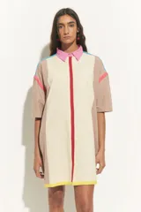Vestido corto tipo túnica color crema con bloques de color beige en hombros y laterales. Presenta cuello camisero rosa, cierre frontal con línea roja y terminación con franja amarilla. Detalles de líneas turquesas y fucsias en hombros.