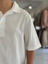 Chomba oversize blanca de lino con mangas cortas, cuello polo y cierre frontal parcial de dos botones. Cuenta con una pequeña etiqueta decorativa en el borde inferior.