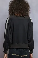Campera negra con diseño strapless, cierre frontal completo y mangas largas con detalle de franjas laterales de encaje. Presenta apliques de tachas metálicas en el cuello y en el ruedo.