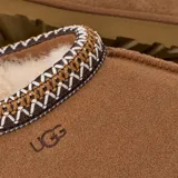 Zuecos UGG modelo Tasman II, confeccionados en gamuza color marrón con forro interno de piel de oveja. Presentan un ribete trenzado decorativo en el cuello y suela de goma resistente.