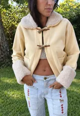 Campera de gamuza color beige con interior y detalles de corderito sintético en cuello y puños. Presenta cierre frontal con botones tipo toggle de madera y cuero, y bolsillos frontales.