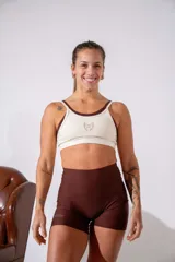 Top deportivo de doble capa en color crema con detalles en marrón oscuro, con breteles finos y logo bordado en el centro del pecho.