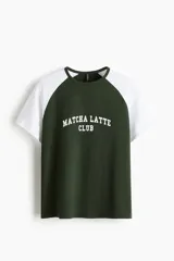 Remera de algodón con mangas raglán cortas en color blanco y cuerpo verde oscuro. Cuenta con cuello redondo ribeteado en verde y un estampado frontal con la inscripción "Matcha Latte Club" en blanco.