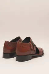 Zapato tipo monk strap con aberturas laterales, confeccionado en cuero vacuno metalizado color bronce. Presenta detalles de perforaciones estilo brogue en la puntera y el talón, y doble hebilla metálica en el empeine.