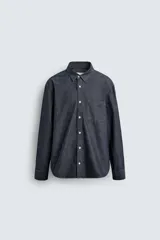 Camisa de corte relajado confeccionada en tejido de algodón denim, con cuello solapa, manga larga con puños abotonados y bolsillo plastrón en el pecho. Presenta cierre frontal de botones y bajo con aberturas laterales.