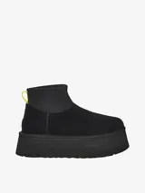 Bota corta tipo botineta de la marca UGG, color negro, con diseño de caña baja y plataforma elevada. Presenta una tira de agarre en color amarillo neón en la parte posterior y una suela de goma gruesa con textura acanalada.