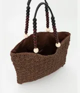 Bolso de mano tipo tote, tejido en punto de ganchillo con textura de conchas, color marrón oscuro. Las asas son de cuero o material sintético oscuro, adornadas con cuentas redondas de madera en tonos marrón oscuro, borgoña y crema.