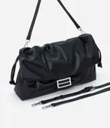 Cartera tipo shoulder bag de tamaño grande, confeccionada en poliuretano con acabado que imita el cuero. Presenta un diseño desestructurado con solapa superior, cierre mediante cordón de ajuste y detalles metálicos plateados en el frente. Incluye correa ajustable para llevar al hombro o cruzada.