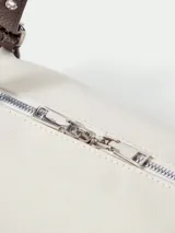 Cartera tipo shoulder bag de diseño cilíndrico, confeccionada en material sintético color crema con textura. Presenta correas largas ajustables en color marrón oscuro con detalles de hebillas y ojalillos metálicos.