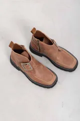 Botines de cuero vacuno color suela con textura envejecida, suela track negra con pespunte en contraste, solapa cruzada con gran hebilla metálica plateada y cierre lateral interior.