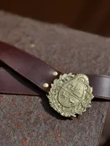 Cinturón de cuero marrón con hebilla metálica grande en forma de escudo nacional.