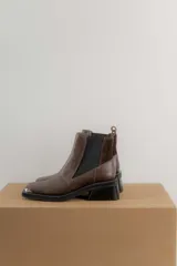 Botas tipo chelsea de cuero color marrón oscuro con acabado brillante. Presentan punta cuadrada con detalle de puntera metálica, elásticos laterales y taco cuadrado bajo.