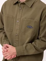 Camisa de gabardina color verde oliva, con cuello clásico, cierre frontal de botones y dos bolsillos frontales con parche. Incluye un pequeño parche con el logo de la marca en el bolsillo izquierdo.