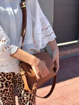 Cartera tipo bandolera de cuero color tostado, con solapa frontal, cierre principal y herrajes metálicos. Incluye dos correas desmontables, una corta para llevar al hombro y una larga ajustable para usar cruzada.