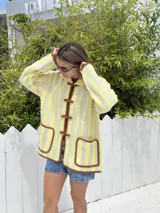 Cárdigan tejido a crochet con hilado liviano color amarillo y beige a rayas verticales. Tiene cuello polo, cierre frontal con nudos y bolsillos frontales con ribete marrón.