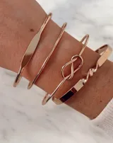 Set de cuatro pulseras esclavas rígidas en tono dorado. Incluye una pulsera con placa central plana, una pulsera fina lisa, una pulsera con nudo infinito y una pulsera con diseño retorcido.